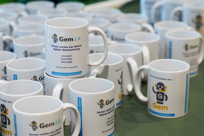Tafel vol witte 'Vrienden van Gem'-mokken met het Gem-logo