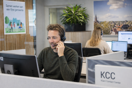 Medewerker van een klantcontactcentrum voert een telefoongesprek achter een beeldscherm, met collega's op de achtergrond