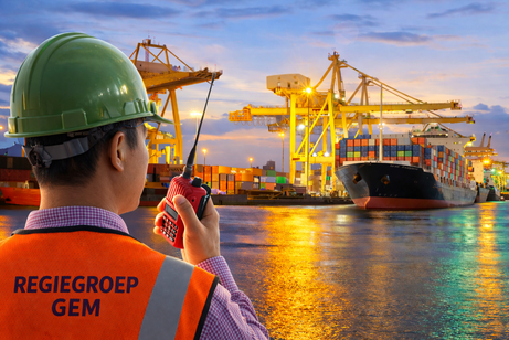 Medewerker met groene helm en oranje hesje met de opdruk 'Regiegroep Gem' kijkt met een portofoon uit over een containerhaven in de avond
